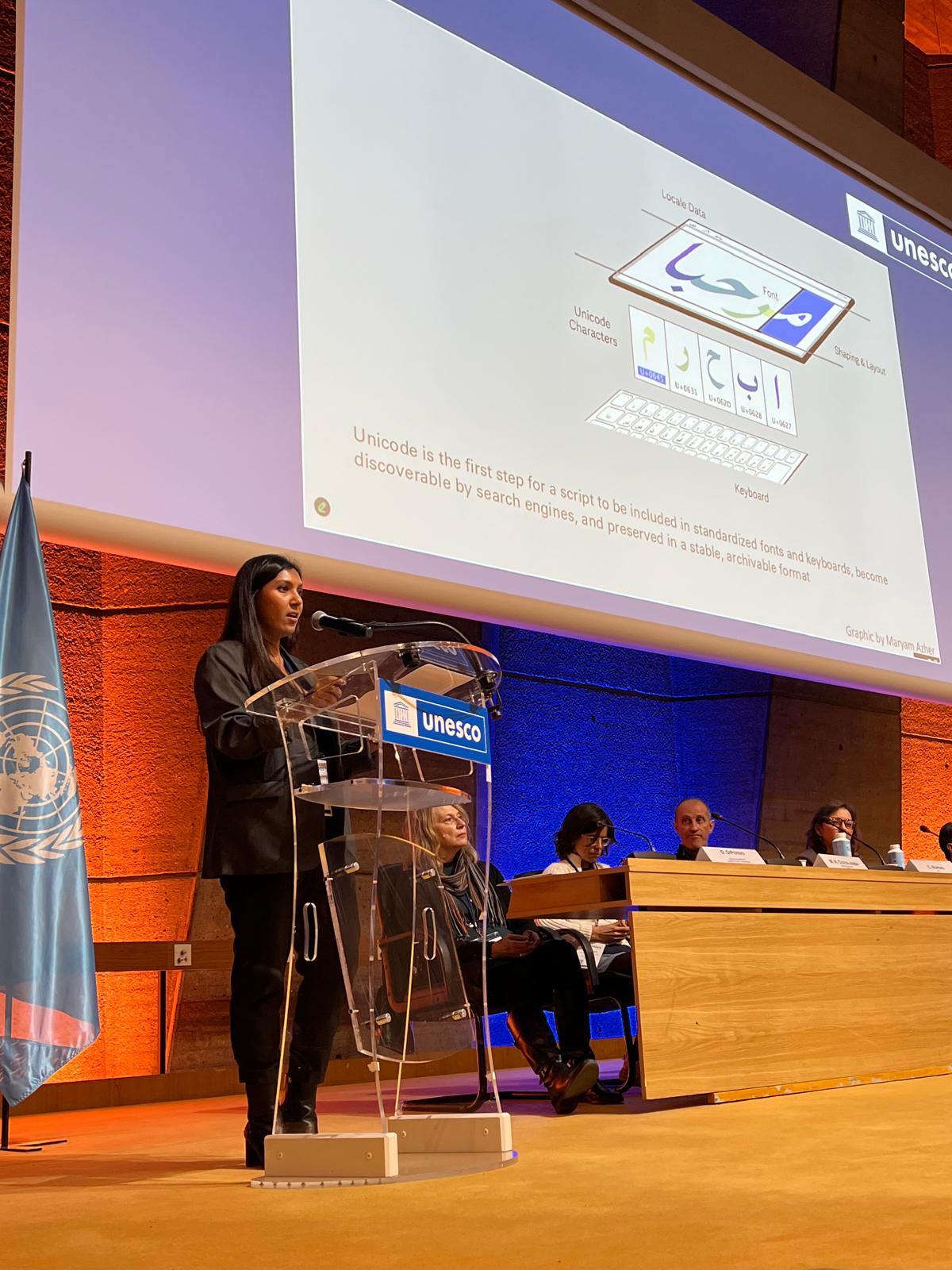 Anushah Hossain standing at a podium at a UNESCO event with diagram of text stack in background