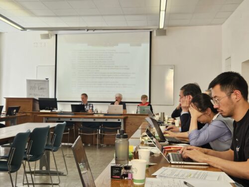 people seated around a table in a classroom, looking at laptops and a projector screen